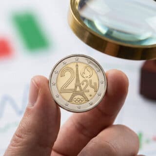 Hand holding a shiny 2 euro Paris 2024 Olympic Games commemorative coin, with a magnifying glass and blurred financial charts.