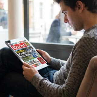 Homme assis lisant le Jornal Record sur une tablette, dans un café. Pâtisseries en arrière-plan.