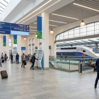 Voyageurs dans le hall lumineux d'une gare moderne avec des panneaux de signalisation et des bornes interactives.